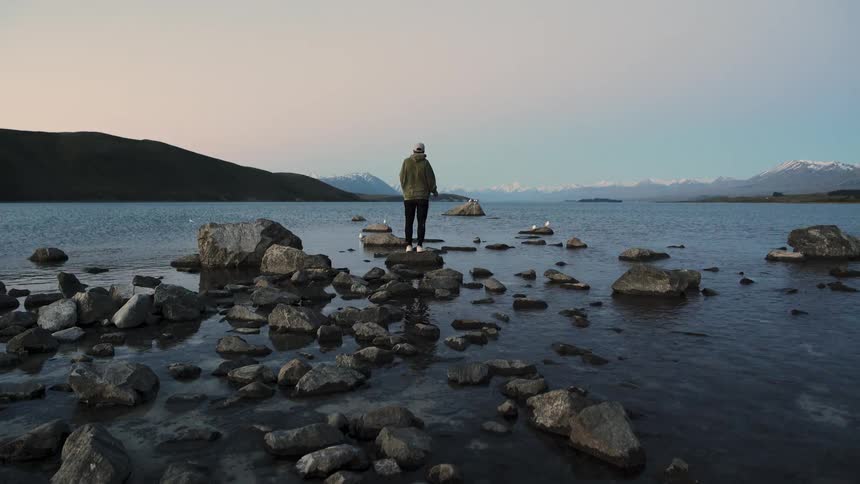 A person walks across rocks to stand in a tranquil lake at twilight, admiring the scenic mountain view