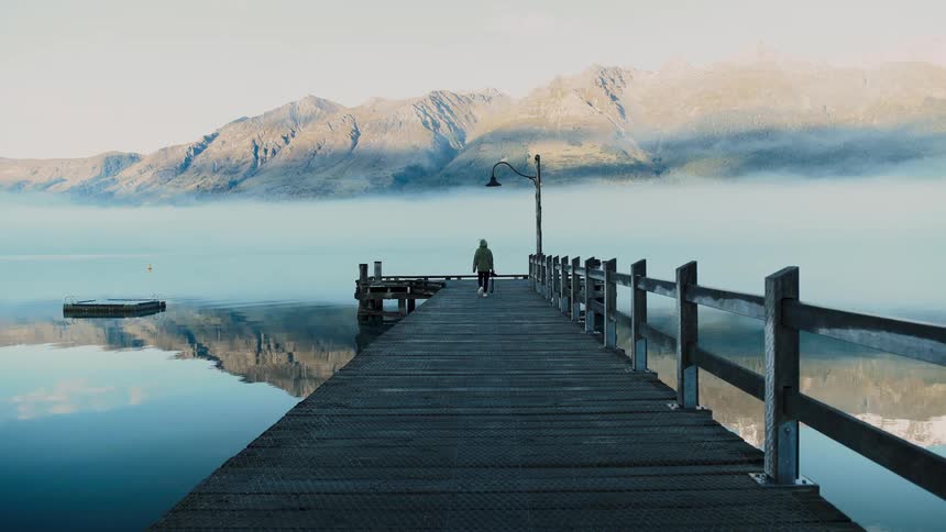 A person wearing a hoodie walks to the end of a wooden pier on a calm, misty lake surrounded by majestic mountains