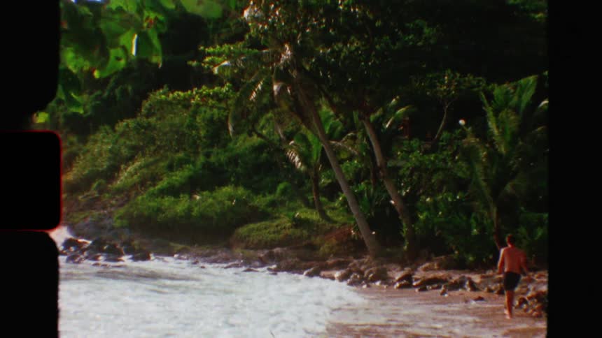 Vintage 8mm film captures a man walking along a secluded tropical beach where lush jungle meets the ocean waves