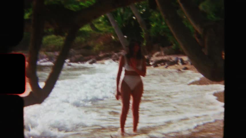 Vintage-style footage shows a woman in a white bikini standing in the shallow waves of a secluded tropical beach