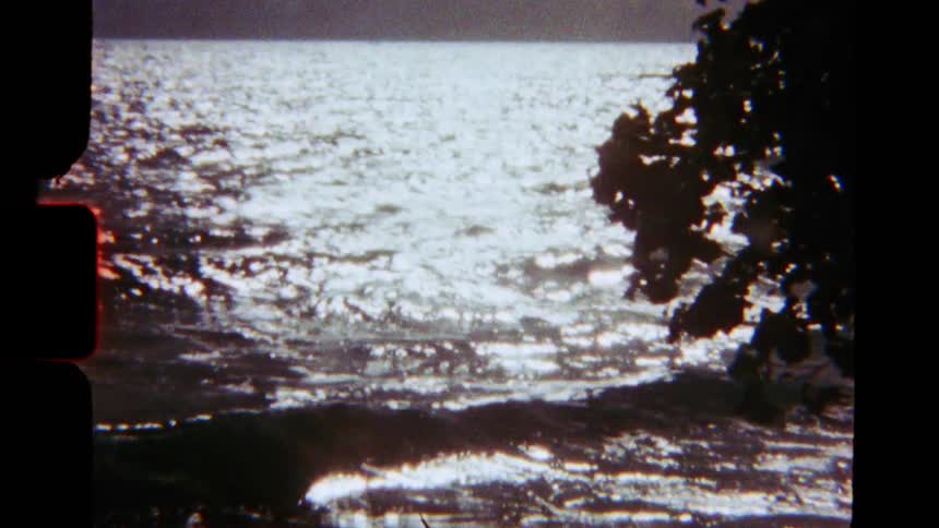 Authentic vintage 8mm footage shows sunlight glittering on the surface of gentle ocean waves under a cloudy sky