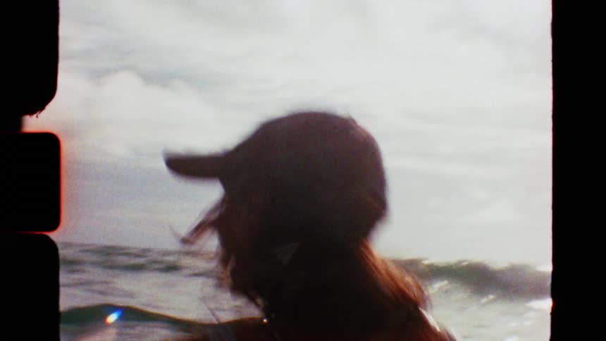 A young woman wearing a backwards cap looks out at the ocean then turns to smile in this vintage 8mm style footage