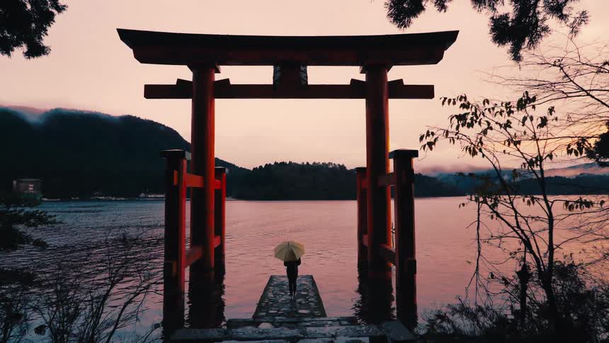 A serene push-in shot captures a lone figure with a yellow umbrella standing on a pier beneath a red Japanese Torii gate in a lake at dusk