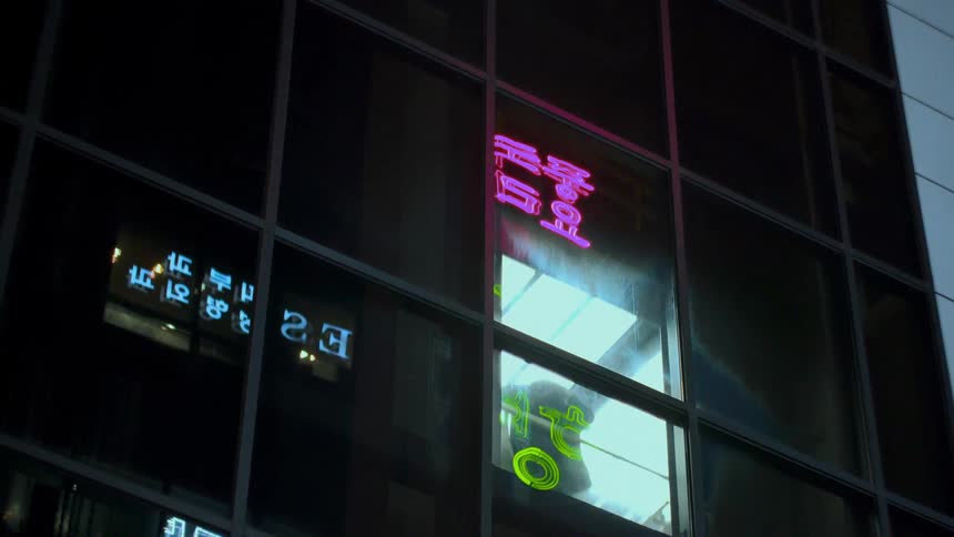 A person rides a glass elevator up a modern building at night, with vibrant neon signs reflected in the windows