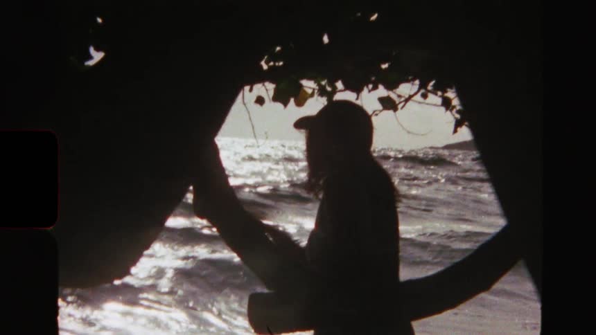 Vintage film captures the silhouette of a woman in a hat relaxing on a tree branch while watching the sunlit ocean waves