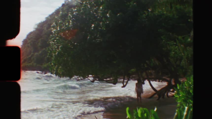 Vintage Super 8 footage reveals a woman wading in the gentle waves of a secluded tropical beach under lush trees