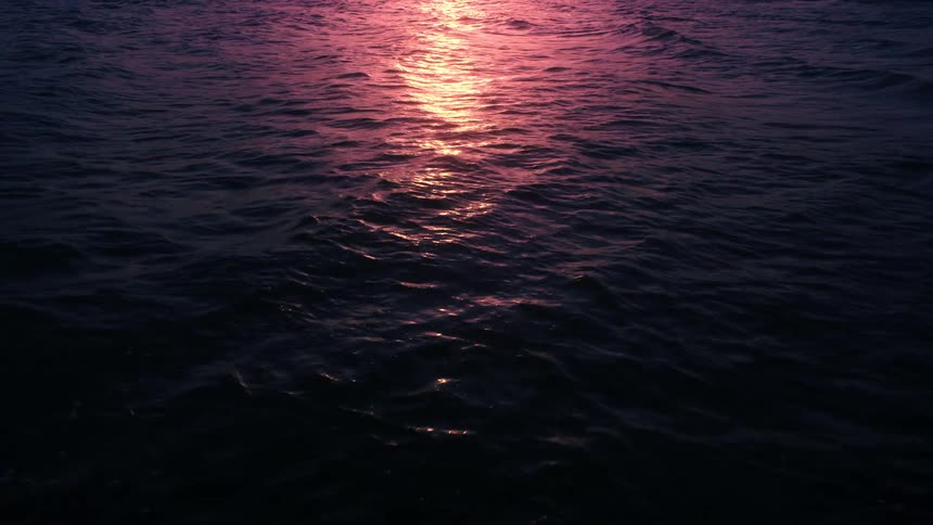 The pink and purple glow of a sunset reflects on the dark, calm ocean waves