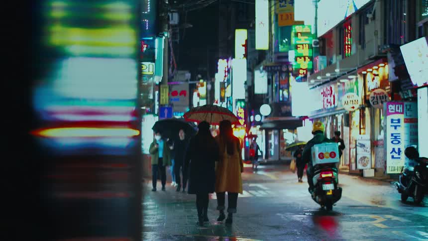 A food delivery driver on a scooter navigates a busy, neon-lit street in Seoul on a rainy night as pedestrians walk by