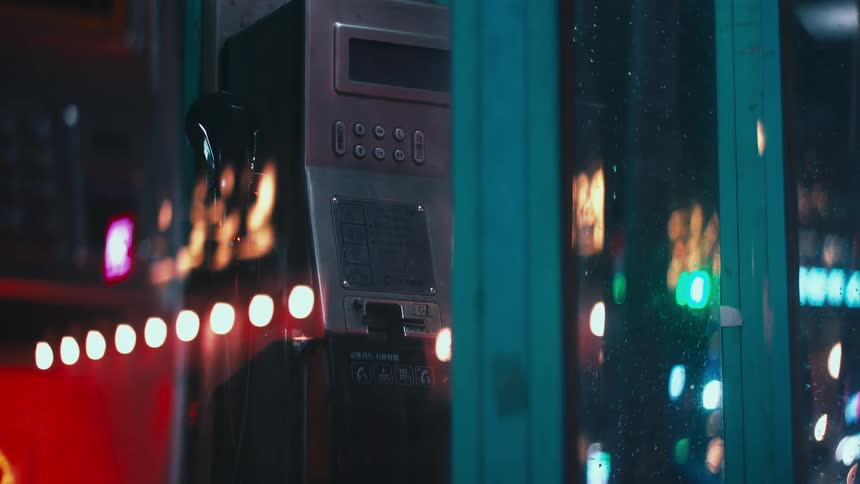 An atmospheric view of a payphone inside a booth on a rainy night, with vibrant city lights reflecting on the wet glass