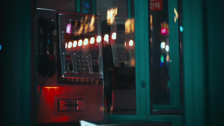 A vintage payphone inside a phone booth at night reflects the colorful, blurry lights of the city