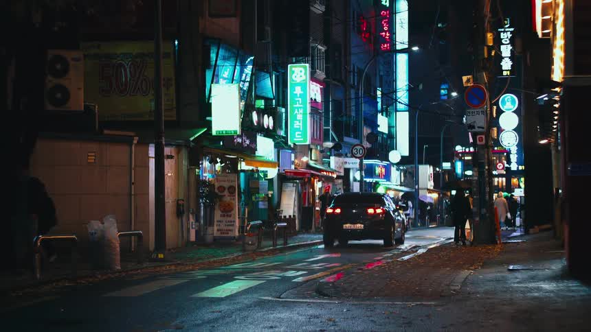 A car drives away down a narrow, neon-lit street in a bustling South Korean city at night