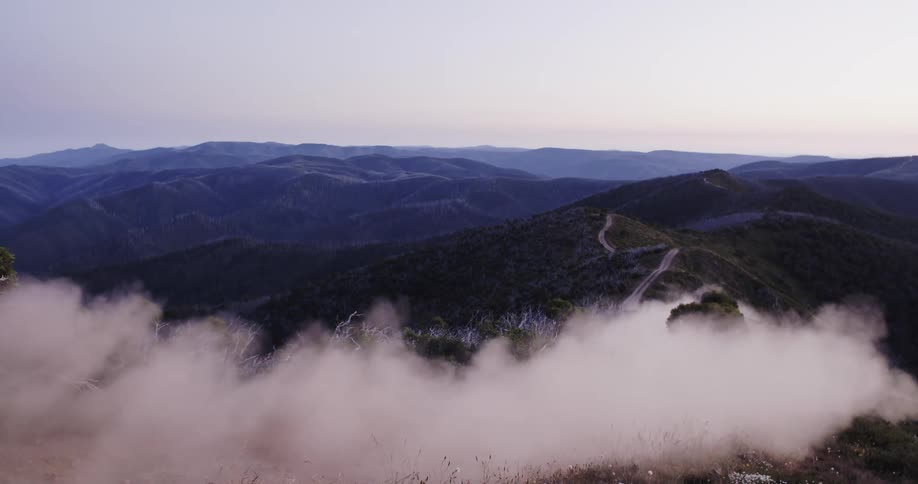 A silver pickup truck drives along a winding dirt road on a mountain ridge at sunset, kicking up a large cloud of dust in slow motion