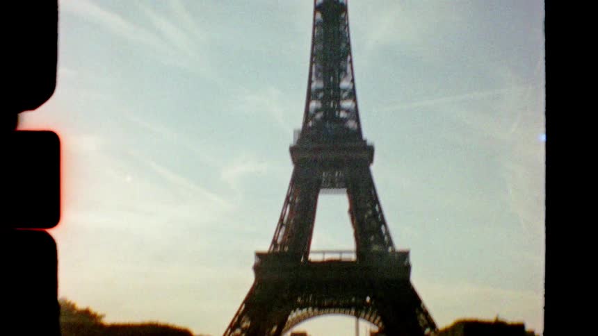 Vintage film footage tilts down from the top of the Eiffel Tower to reveal tourists relaxing on the lawn below on a sunny day in Paris