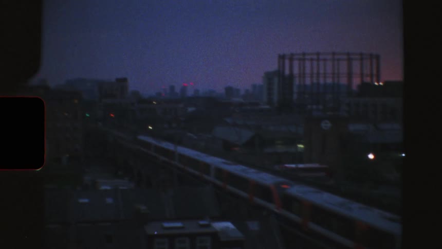 Vintage 8mm film footage captures the London city skyline at dusk as a commuter train passes by in the foreground