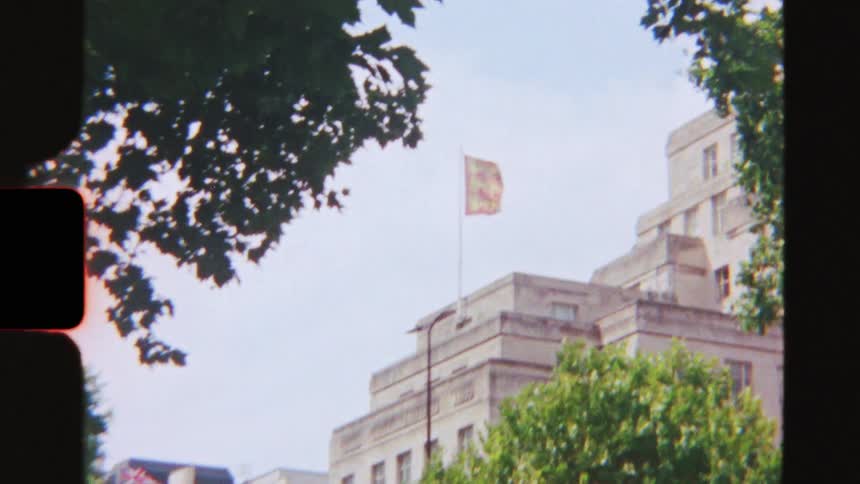 Handheld vintage 8mm footage slowly zooms in on the Royal Standard flag waving above a building in London