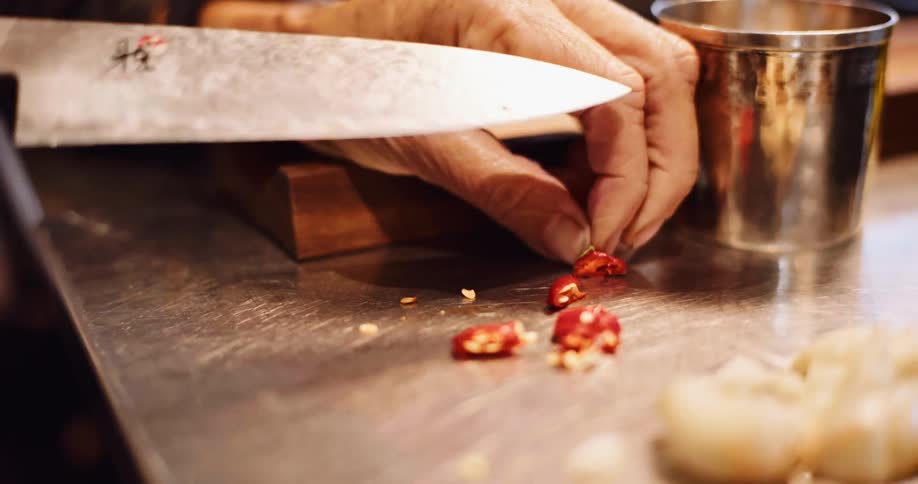 A chef's hands carefully slice a red chili pepper with a beautiful Japanese Damascus steel knife on a stainless steel counter