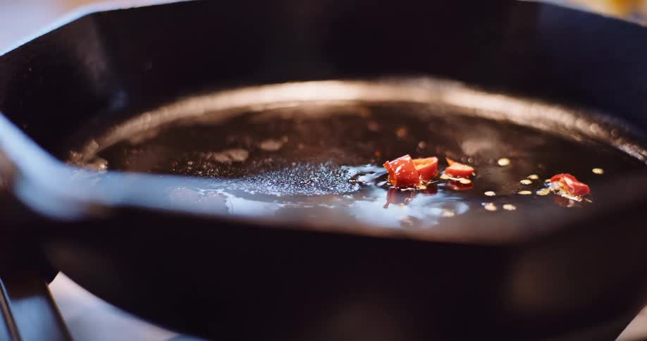 A close-up slow-motion shot of red chili peppers and seeds sizzling in hot oil in a cast iron frying pan