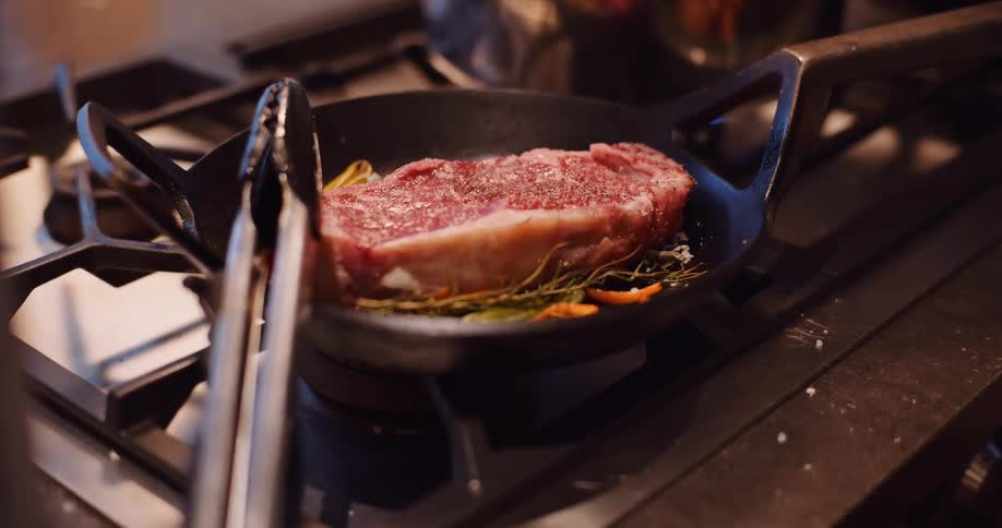 A seasoned raw steak begins to cook in a cast iron skillet with herbs over a gas flame