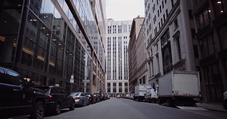 A low-angle establishing shot of an empty city street canyon flanked by tall buildings and lined with parked vehicles
