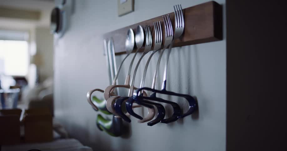 A collection of adaptive utensils with large handles hang neatly on a wooden wall-mounted rack inside a home