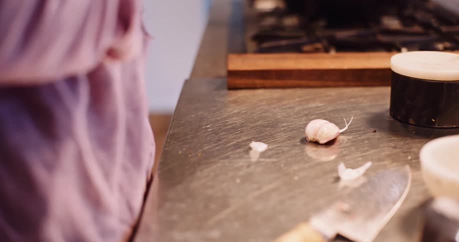 A close-up slow-motion shot of an elderly person's veiny hands peeling a clove of garlic on a stainless steel kitchen counter