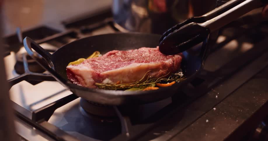 A chef carefully places a seasoned raw steak into a hot skillet with herbs to sear in slow motion