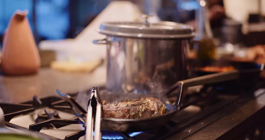 A seasoned steak sizzles and steams in a hot pan on a gas stove in a busy kitchen