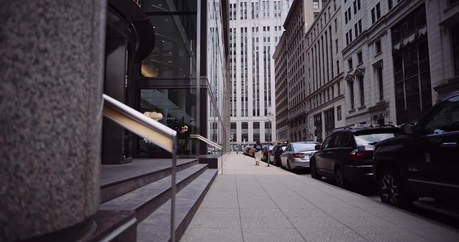 A woman carrying a box and tote bag walks away from the camera down a city sidewalk lined with parked cars and tall buildings