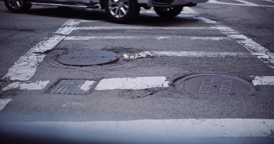 A low-angle view shows the feet of pedestrians walking across a weathered city crosswalk in Boston