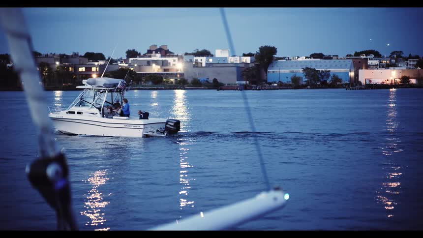 A fishing boat slowly cruises across the water at dusk as the city lights reflect on the calm surface