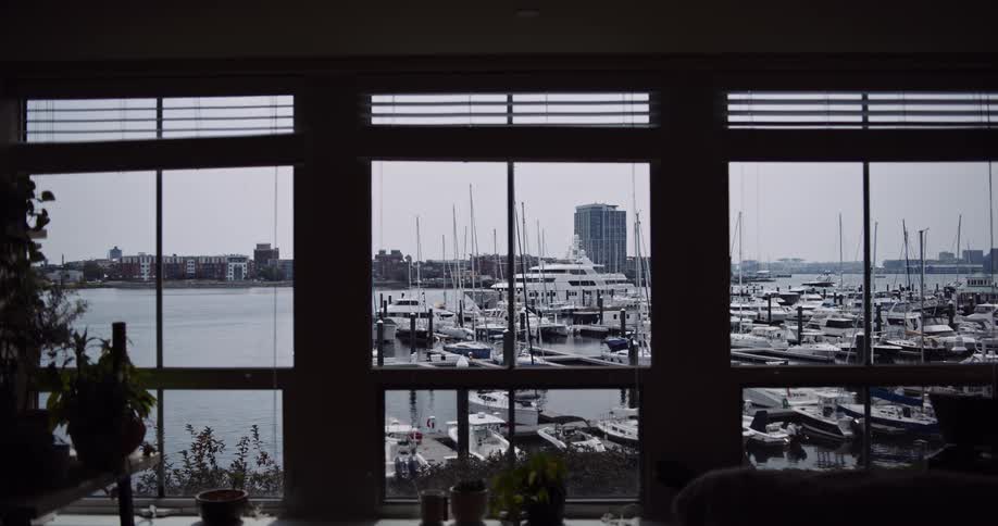 A serene view of a bustling city marina filled with boats and yachts, seen from the window of a dark, silhouetted apartment