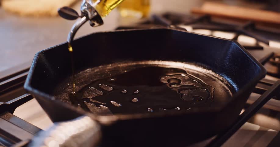 A person pours golden olive oil from a dispenser into a black cast iron skillet in slow motion