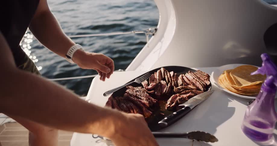 A person slices freshly grilled steak on the deck of a boat, with a plate of tortillas nearby and the sparkling ocean in the background