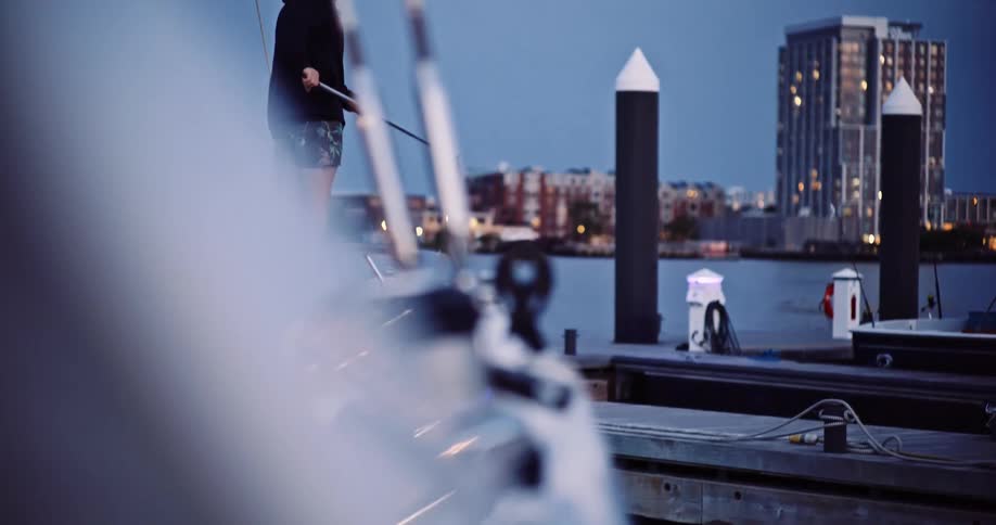 A man in a hoodie stands on a boat looking out at the city from a marina at dusk