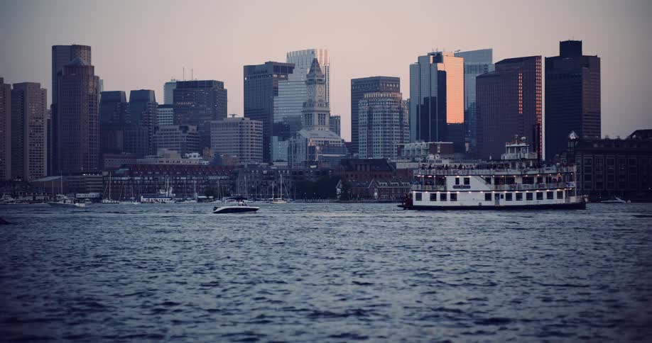 A scenic view of the Boston city skyline from across the harbor at dusk as a large tour boat sails by