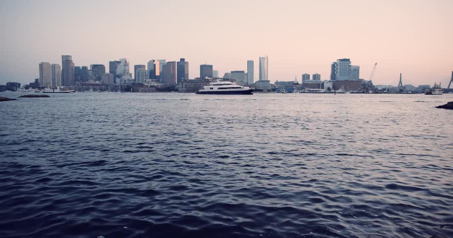 A wide, static shot of the Boston city skyline from across the harbor as a ferry sails past during a hazy sunset