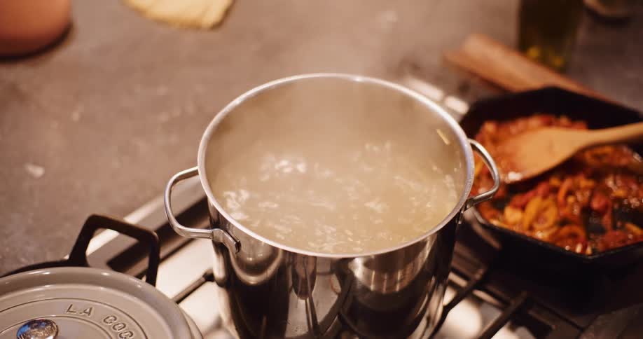 A person stirs long noodles with tongs in a pot of boiling water on a gas stovetop