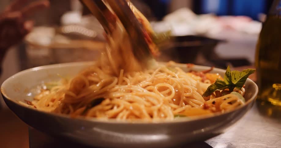 A chef uses tongs to toss a steaming bowl of fresh spaghetti with tomato sauce and basil