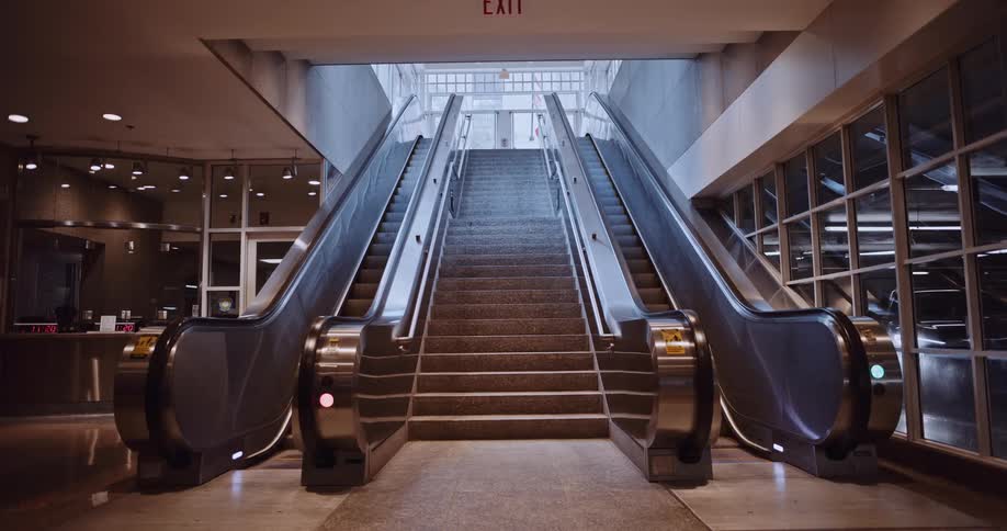 A symmetrical shot of a stationary escalator and staircase leading up towards a brightly lit exit in a modern building
