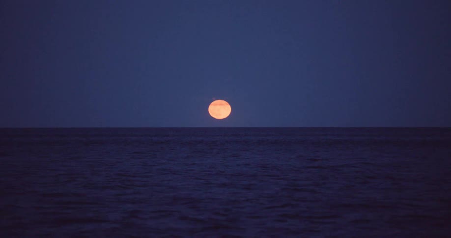 A serene view of an orange full moon rising over the dark, calm ocean at night