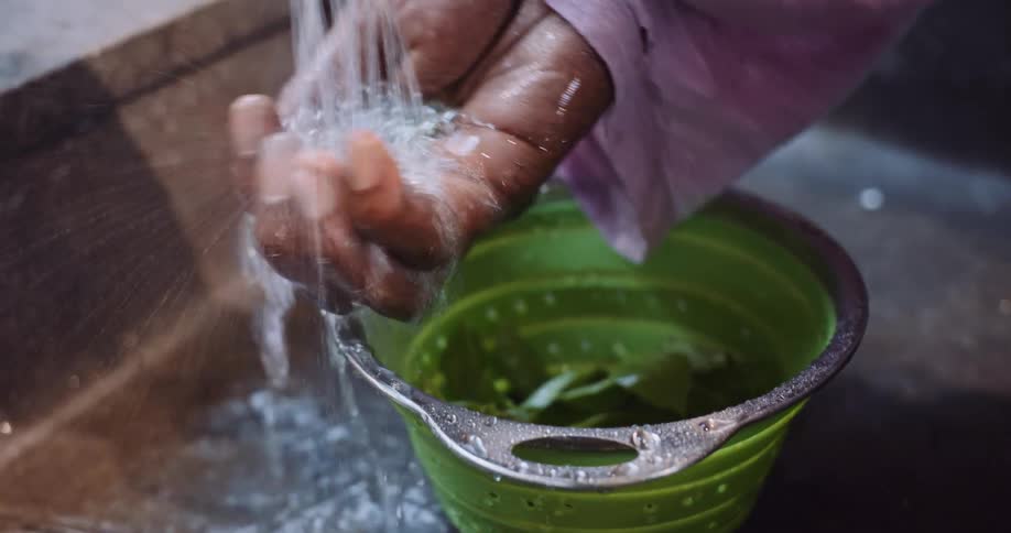 A close-up slow-motion shot of a person's hands washing fresh basil leaves in a green colander under running water
