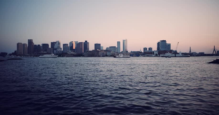A tranquil, wide shot of the Boston skyline across the rippling harbor waters during a beautiful sunset