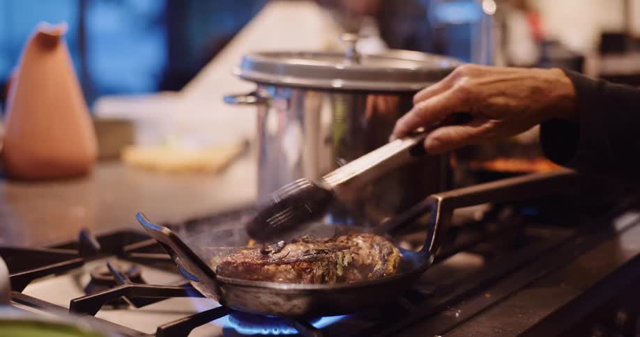 Close-up of a chef using tongs to flip a seasoned steak as it sizzles in a hot pan on a gas stove in slow motion