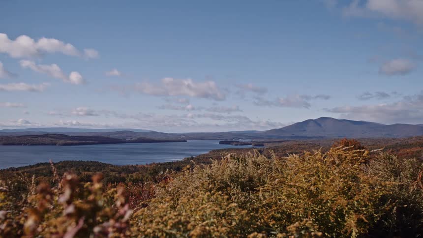 A serene landscape of a vast lake nestled between rolling hills and mountains during peak autumn season on a sunny day