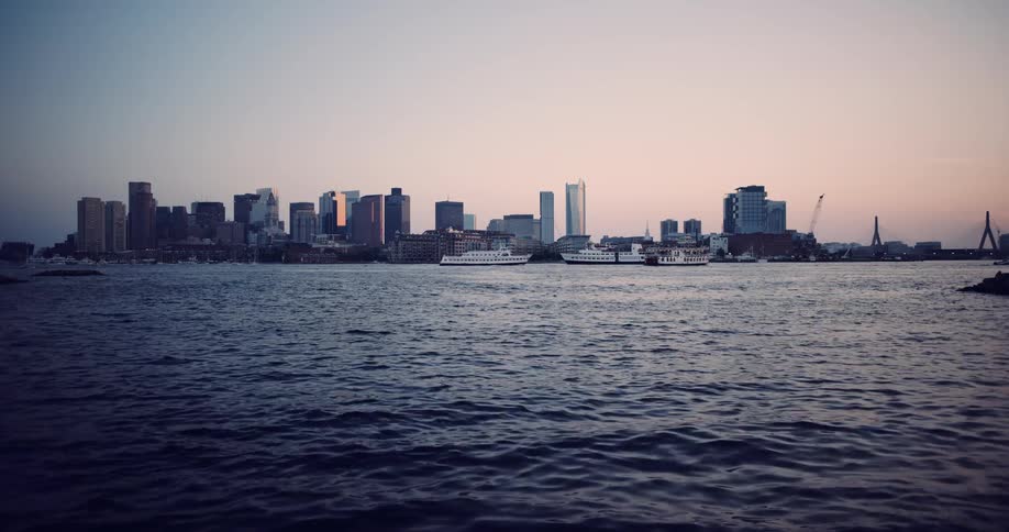 The picturesque Boston city skyline is silhouetted against a soft sunset sky as boats travel across the calm harbor