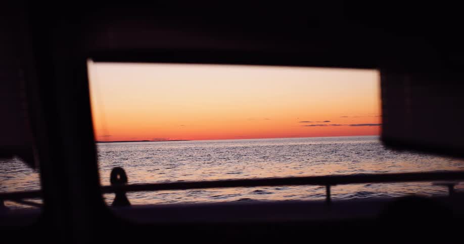A calm ocean sunset with a vibrant orange sky is seen through the rectangular window of a boat cabin