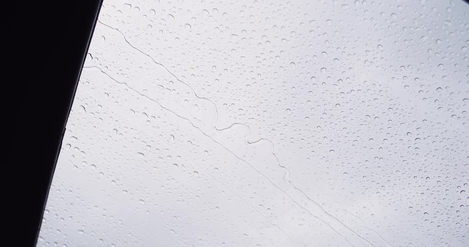 Raindrops slowly trickle down a window pane against a bright, overcast sky