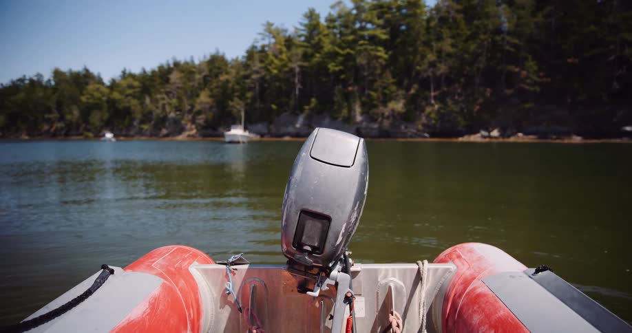 A point-of-view shot from the back of a red inflatable dinghy with an outboard motor moving away from a lush, tree-covered shoreline on a sunny day