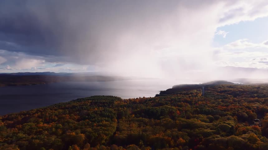 A dramatic rainstorm moves across a lake as sunlight breaks through the clouds over a vast autumn forest