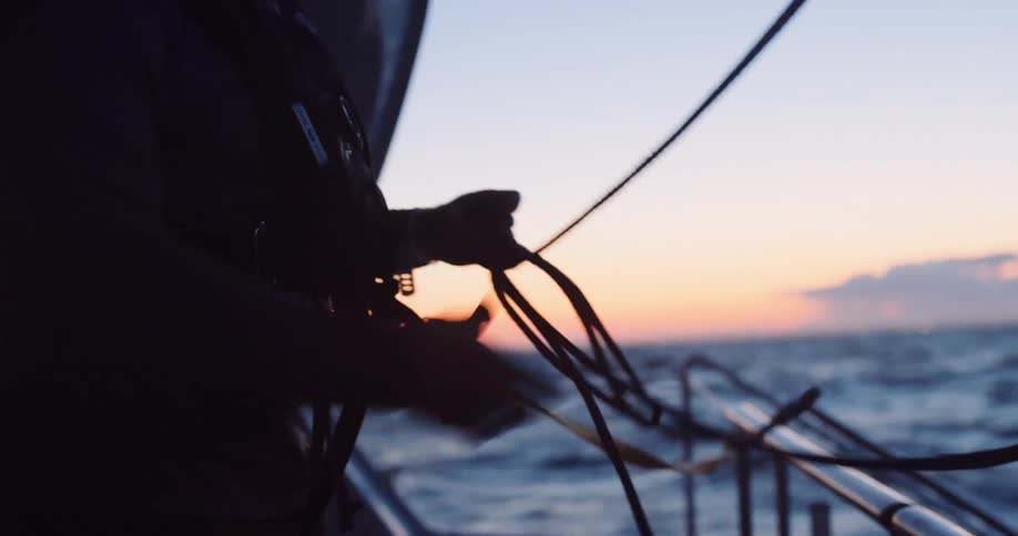 A sailor wearing a life vest carefully coils a rope on the deck of a sailboat during a stunning sunset over the open ocean